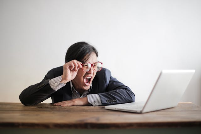 Frustrated man in a suit yelling at his laptop while adjusting his glasses, sitting at a wooden table.